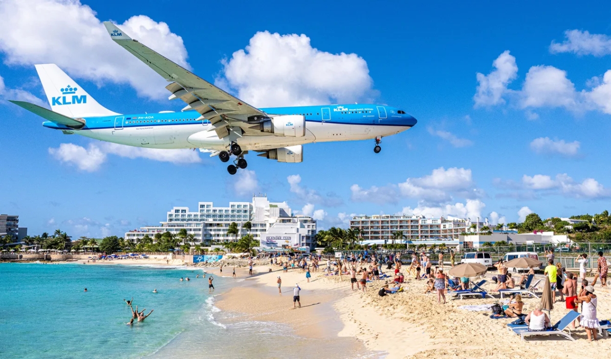 A KLM Royal Dutch Airlines flight preparing to land at Princess Juliana International Airport, hovering spectacularly low over beachgoers at Maho Beach, a globally recognized plane-spotting destination uniquely situated where the runway touchdown zone sits just feet away from the public beach, on the Caribbean island of Sint Maarten.