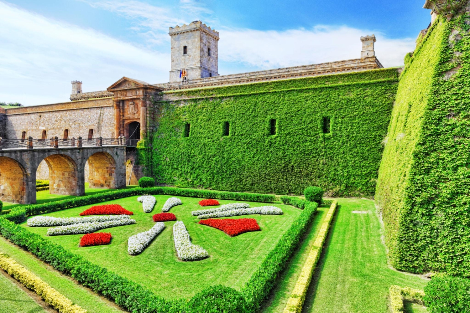 Montjuic Castle in Barcelona, Spain, featuring its iconic ivy-covered stone walls and a meticulously landscaped flower garden in the moat.