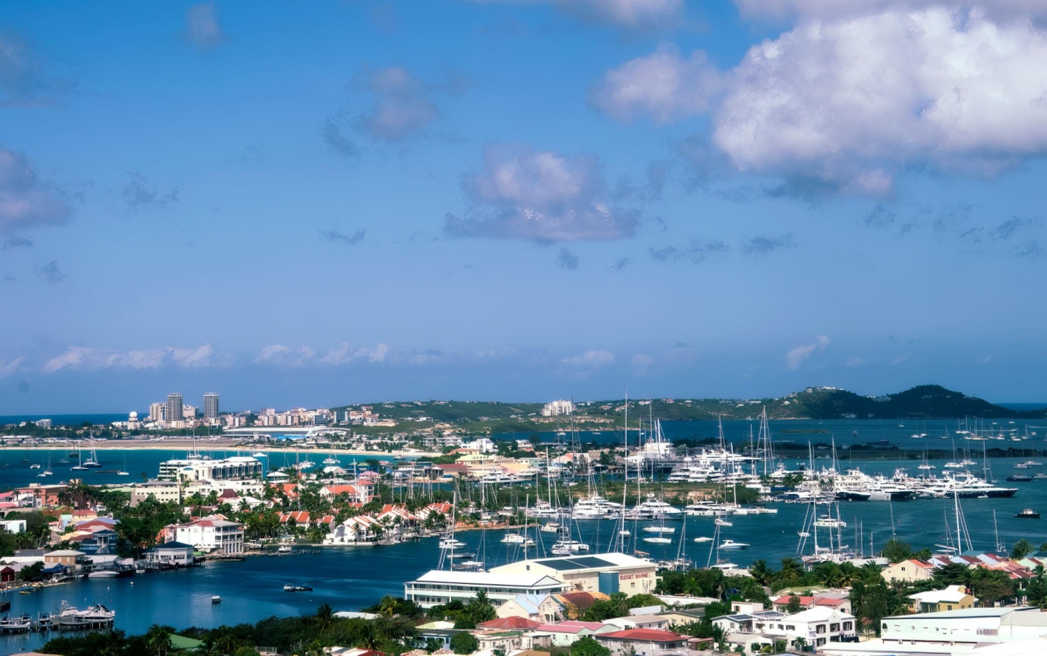 Simpson Bay Lagoon on the island of Sint Maarten in the Caribbean featuring a crowded marina filled with numerous sailboats and luxury yachts, and the town of Simpson Bay with white-roofed buildings and resorts bordering the blue water.